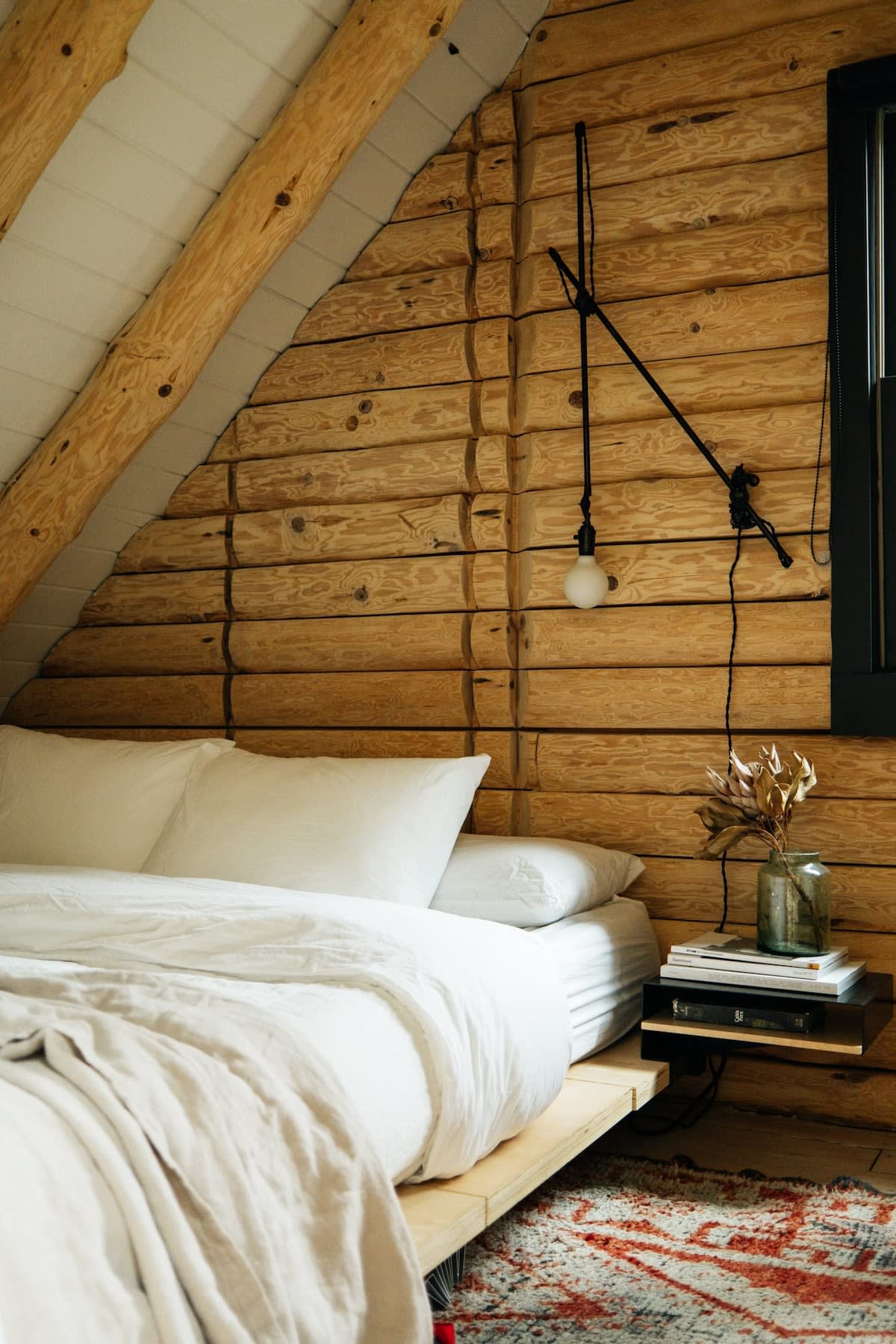 Cozy bedroom with a sloped white ceiling and rustic log walls, featuring a bed with white linens. A black swing-arm wall lamp provides lighting, and a small floating nightstand holds books and a vase with dried flowers.