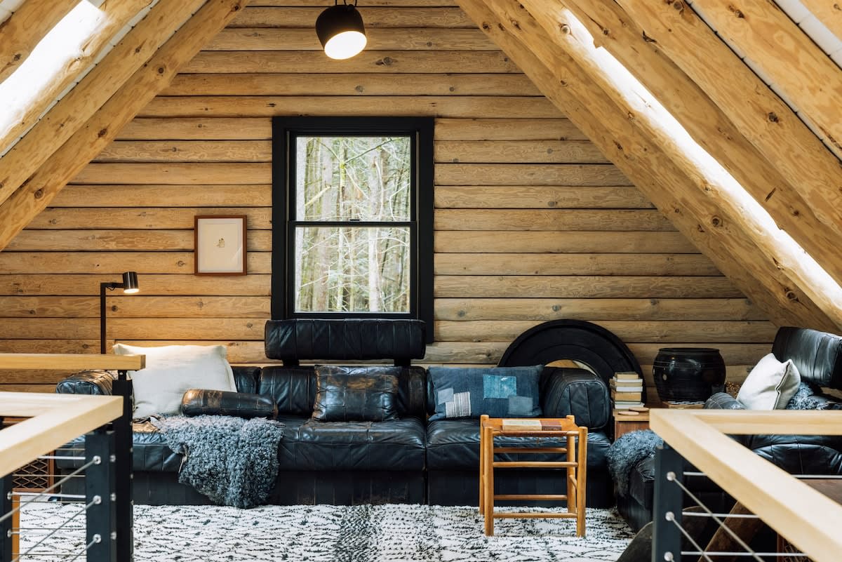A cozy loft living area features a large black leather sectional sofa with throw pillows, a patterned rug, and natural log walls and sloped wooden ceilings, with a window overlooking the forest.