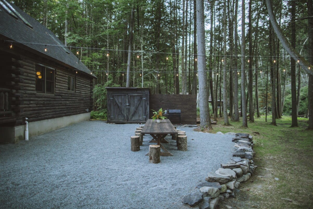 Outdoor dining area with a long wooden table and tree stump seats on a gravel surface, bordered by a low stone wall. String lights hang overhead, and a dense forest surrounds the area.