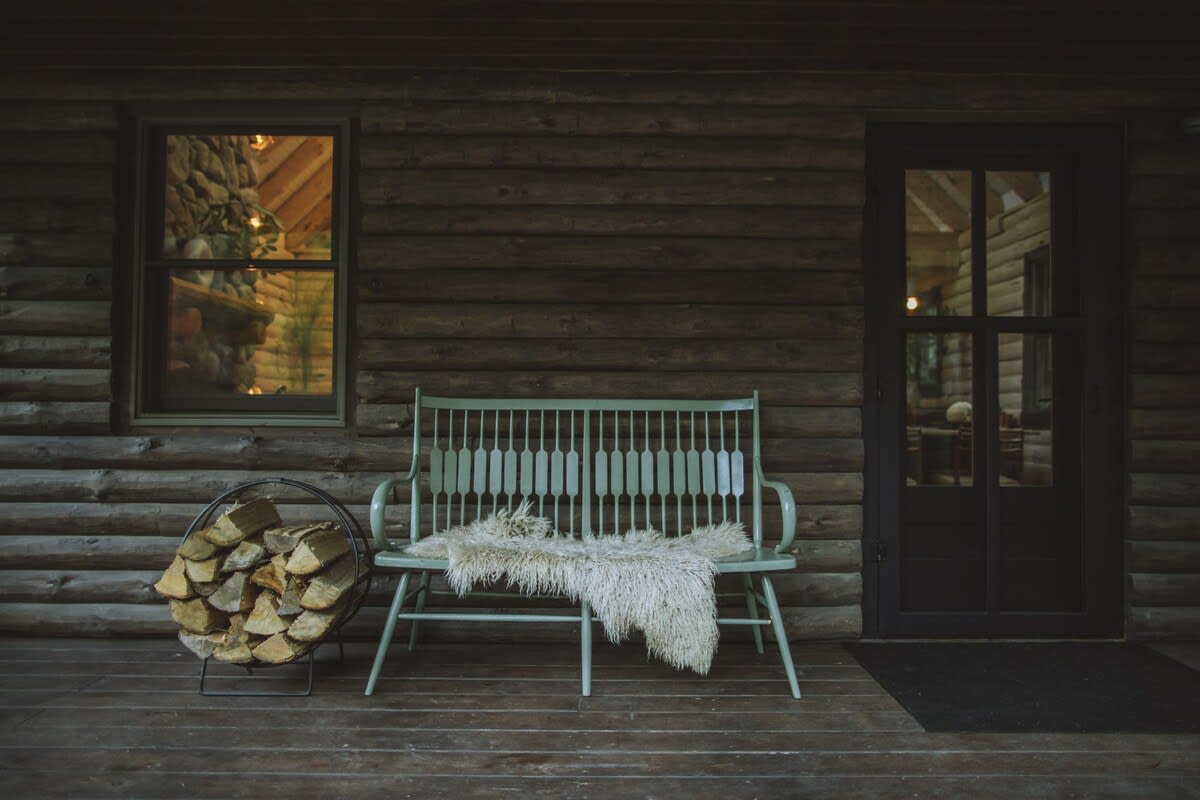 Detail of the log cabin's porch with a light green wooden bench adorned with a white faux fur throw, next to a metal log holder filled with firewood. A window and a dark door are visible.