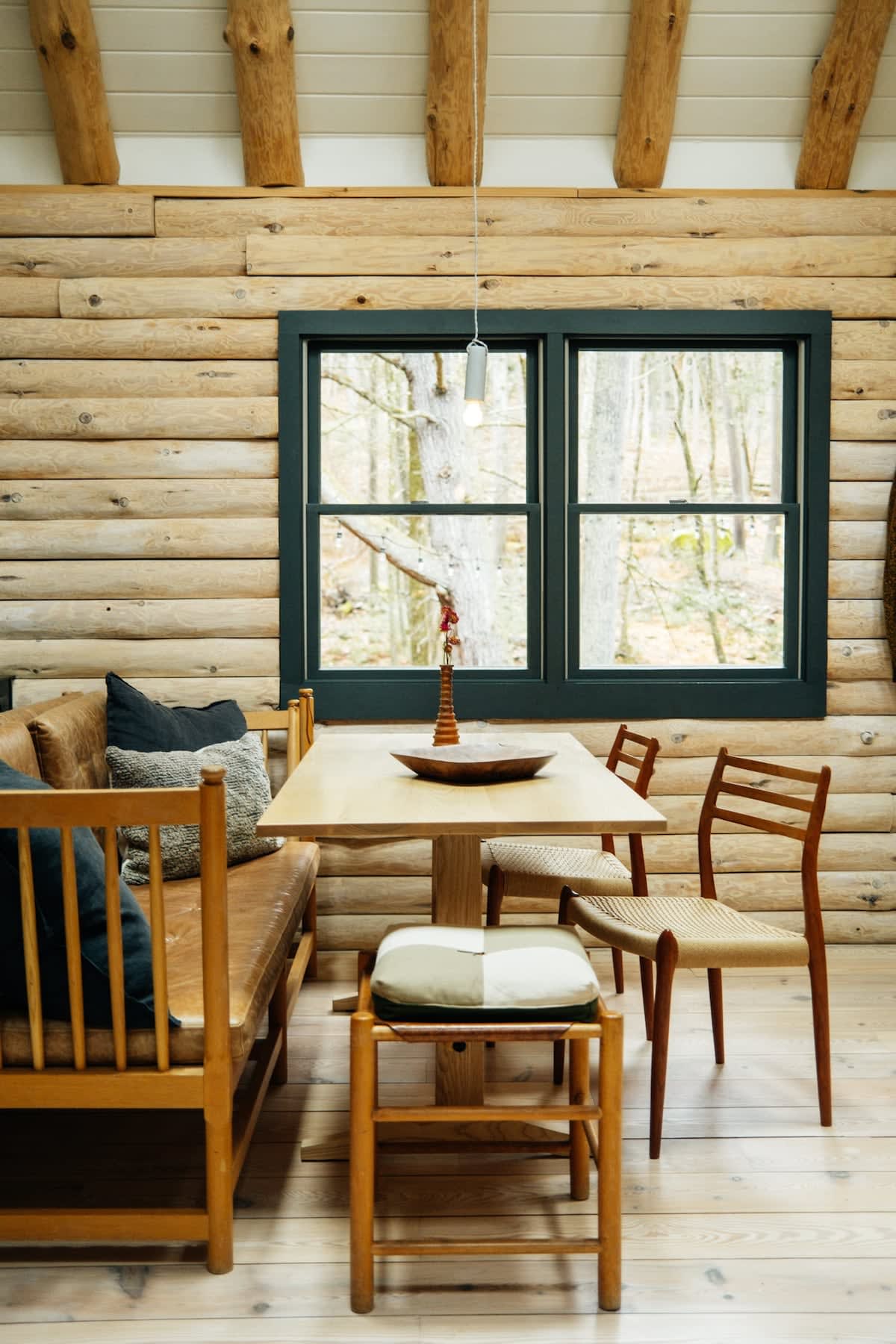 A close-up of the dining area shows a light wood table with a built-in leather bench and two wooden chairs with woven seats, positioned against a log wall with a dark-framed window overlooking a wooded area. A small wooden stool with a cushion is also present.