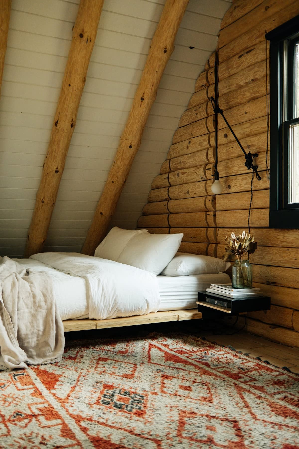 Rustic bedroom with a sloped ceiling, exposed log beams, and log walls, featuring a low-profile wooden bed with white bedding. An orange and white patterned rug covers the floor, and a black-framed window is visible on the right.
