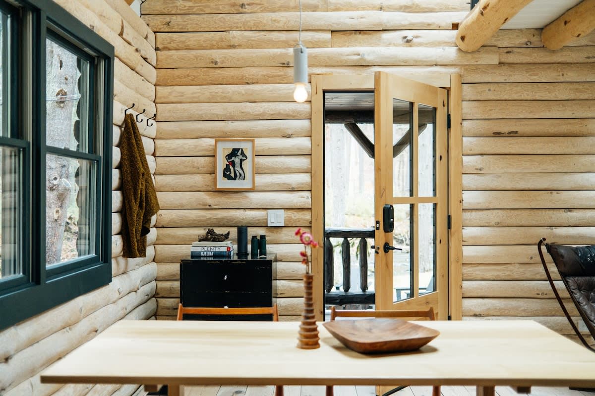 A cozy dining area within the log cabin, featuring light-colored log walls and a light wooden dining table with matching chairs. A black cabinet with decor, a window with dark trim, and an open door leading to the porch enhance the rustic charm.