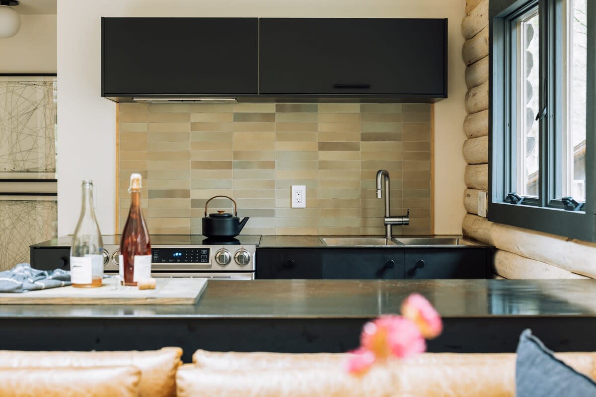 A modern kitchen area features dark flat-panel cabinetry, a light-colored subway tile backsplash, and stainless steel appliances including a range and sink. A window looks out to the exterior, and a kettle sits on the stovetop.