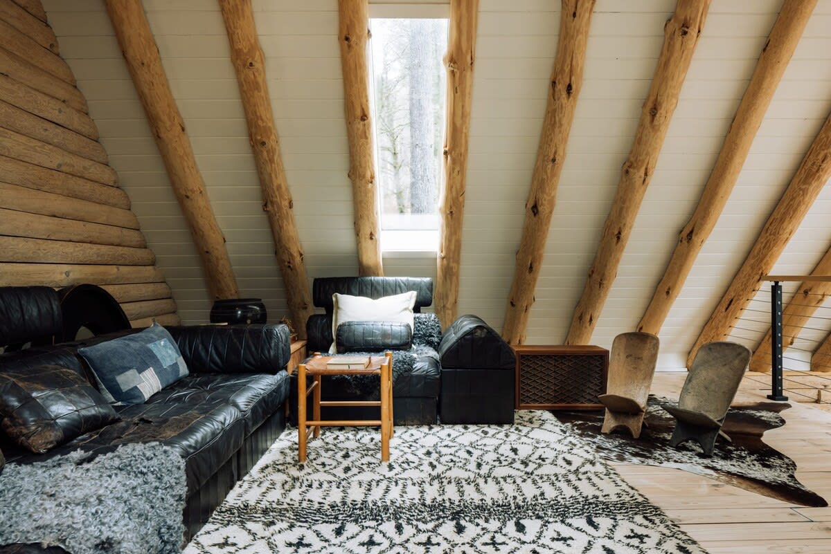 A cozy loft living area features a black leather sofa and armchair, a small wooden side table, and a large patterned rug. Exposed wooden beams and log walls create a rustic aesthetic, with a tall window providing natural light.