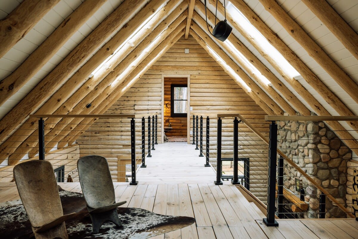 Wide-angle view of a rustic loft or hallway with exposed log beams and light wood plank flooring. A black metal railing with wire infill overlooks the lower level, and a stone wall is visible on the right. Two carved wooden chairs sit on a cowhide rug.