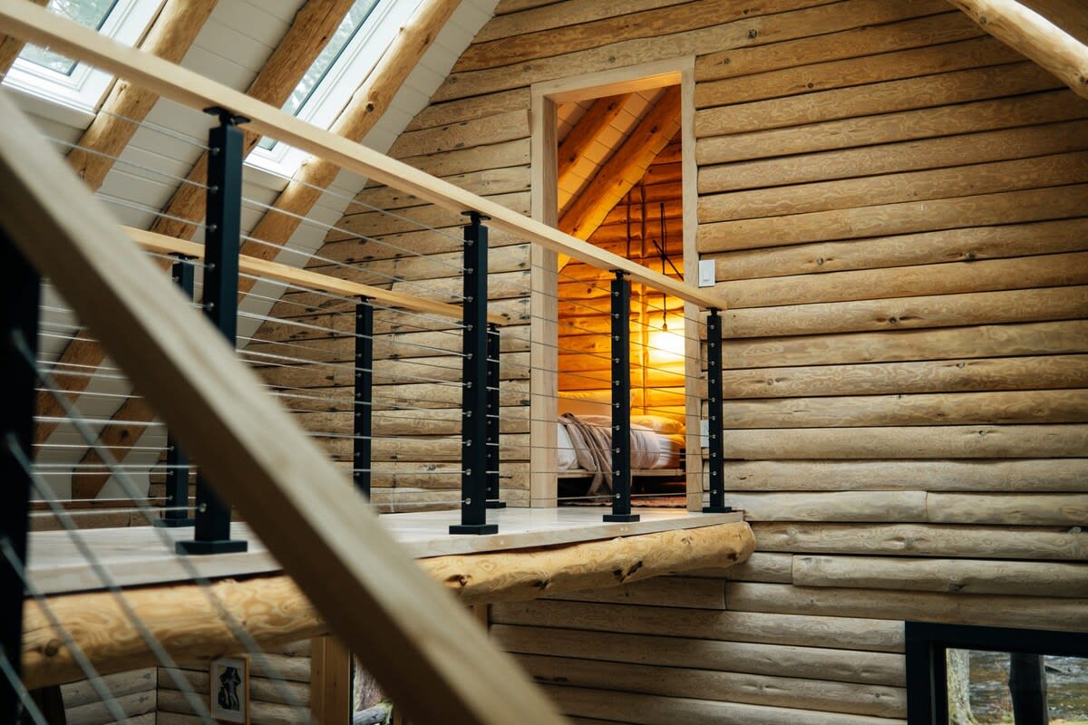 A view from a loft area shows a wooden railing with black metal spindles, looking towards a doorway leading into another room with warm lighting. The space features log walls and exposed wooden beams, characteristic of a cabin.