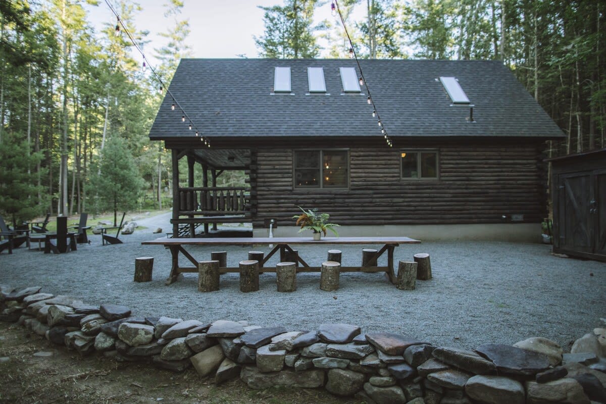 Side view of the log cabin showing a long outdoor dining table with tree stump seats on a gravel patio, illuminated by string lights. The cabin has skylights and is surrounded by tall trees.