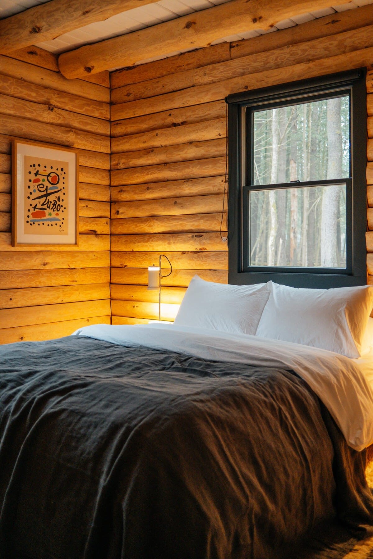 Cozy bedroom featuring log-style wood plank walls and a large window looking out to the forest. A bed with white and dark grey bedding is illuminated by a bedside lamp, with abstract art on the wall.