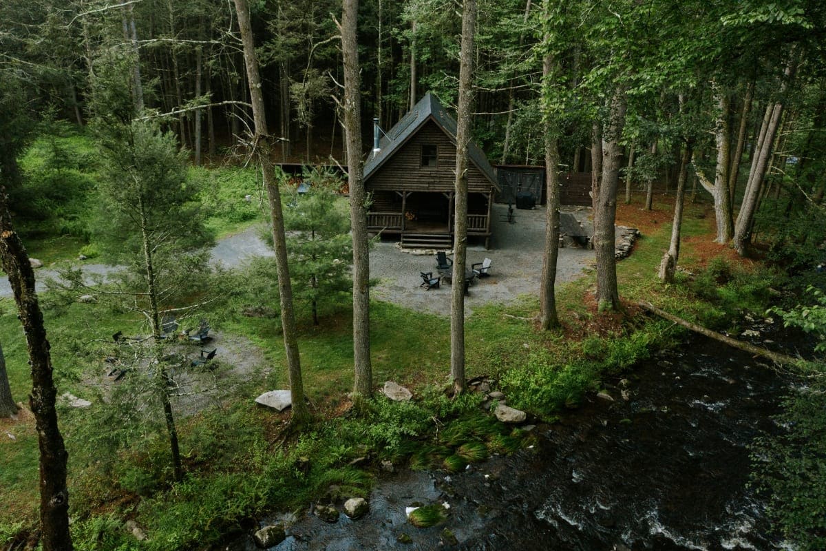 Aerial view of a rustic log cabin nestled in a dense green forest, with a dark river flowing alongside. Outdoor seating areas are visible on the gravel path.