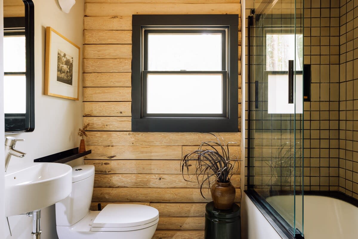 Modern bathroom with horizontal log walls and a white toilet and sink. A black-framed window is centered above the toilet, and a glass shower enclosure is visible on the right.