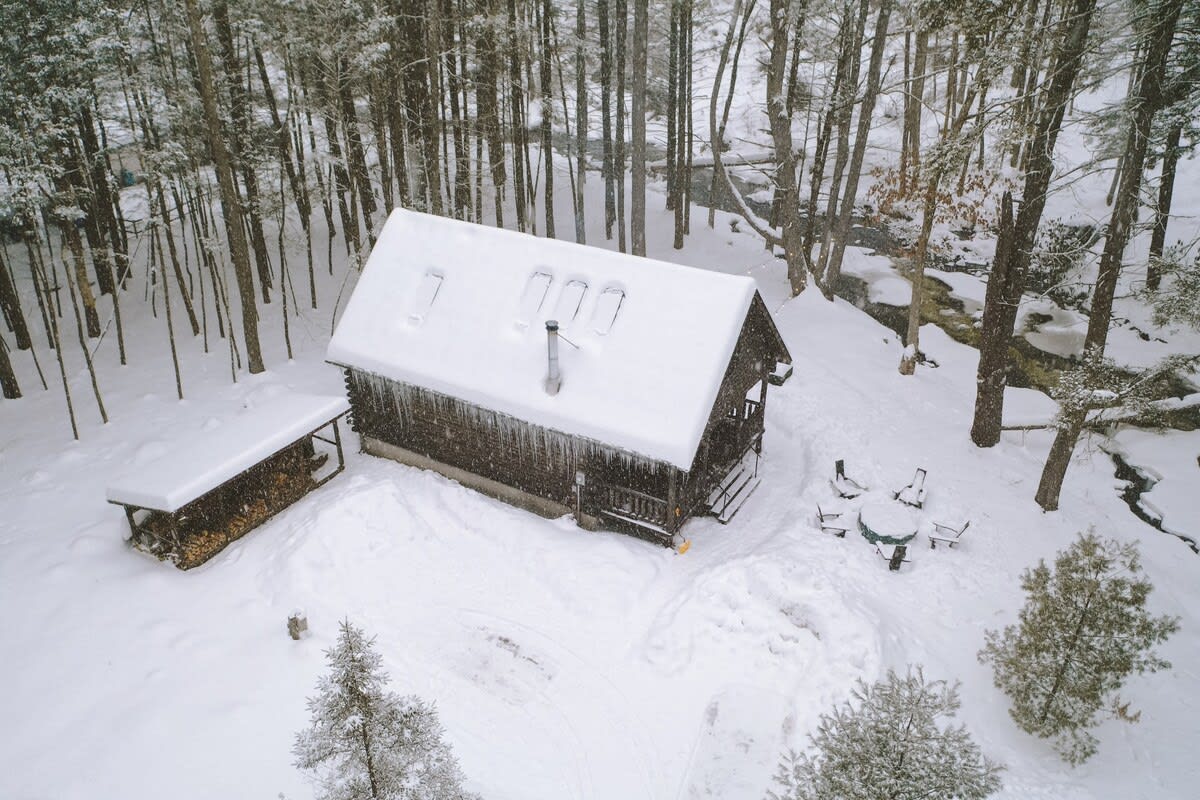 Aerial view of the snow-covered log cabin with a prominent chimney and skylights, surrounded by a pristine white landscape. A wood storage shed is visible, and a partially frozen river flows nearby.
