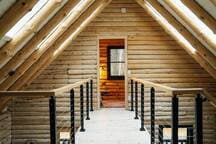 A narrow loft walkway with light wooden floors and sleek black metal railings, offering a view over the main living space. The walls are constructed from light-colored logs, complemented by exposed log beams supporting the vaulted ceiling.