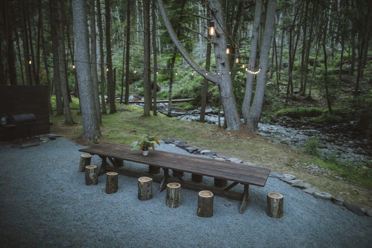 A long, rustic wooden dining table with tree stump seating is set on a gravel patio, surrounded by tall trees and overlooking a flowing river, with string lights overhead.