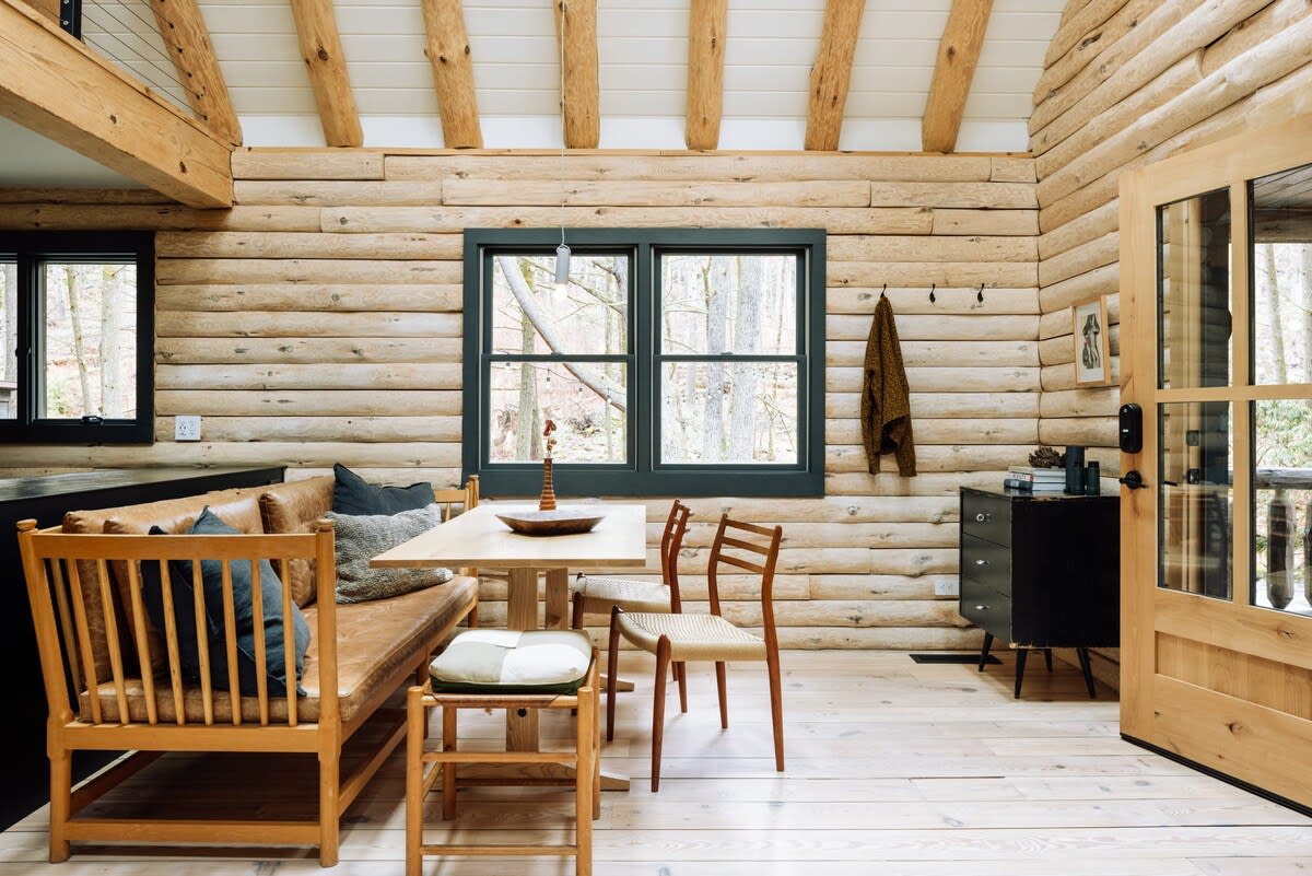 A cozy dining area in a log cabin features a light wood table with a built-in leather bench and two wooden chairs, set against a log wall with a dark-framed window looking out to trees. A dark wood cabinet with drawers and a wooden door are also visible.