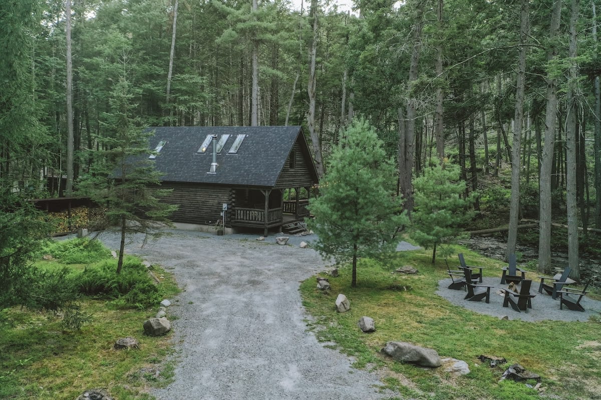 Elevated view of a dark wood log cabin with a covered porch and a dark shingled roof, surrounded by tall green trees. A gravel driveway leads to the cabin, with an outdoor seating area to the right.