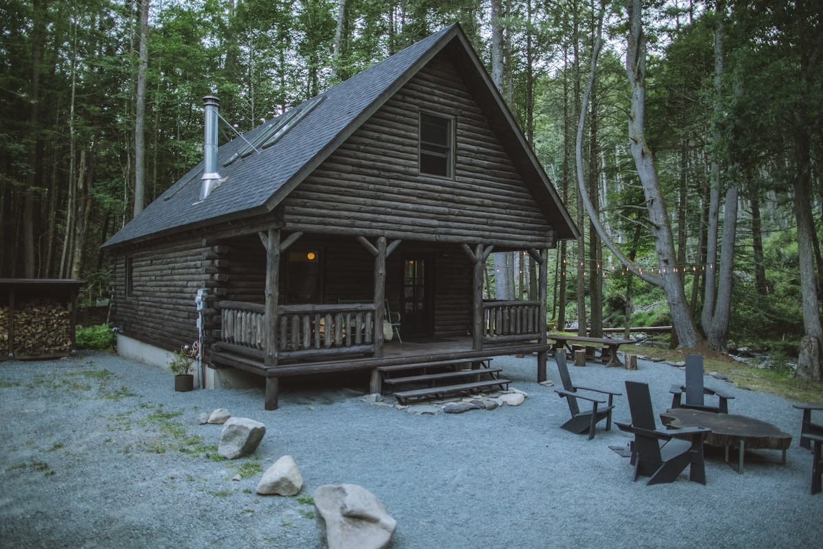 A rustic log cabin with a dark gray roof and a welcoming front porch, nestled among tall green trees. The gravel yard features outdoor seating and a fire pit area, creating a serene forest retreat.