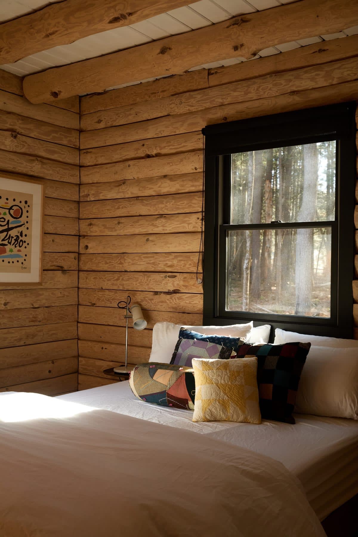 Sunlit corner of a bedroom with log walls and a bed adorned with colorful throw pillows. A black-framed window offers a glimpse of the surrounding forest, and a small lamp sits on a bedside table.