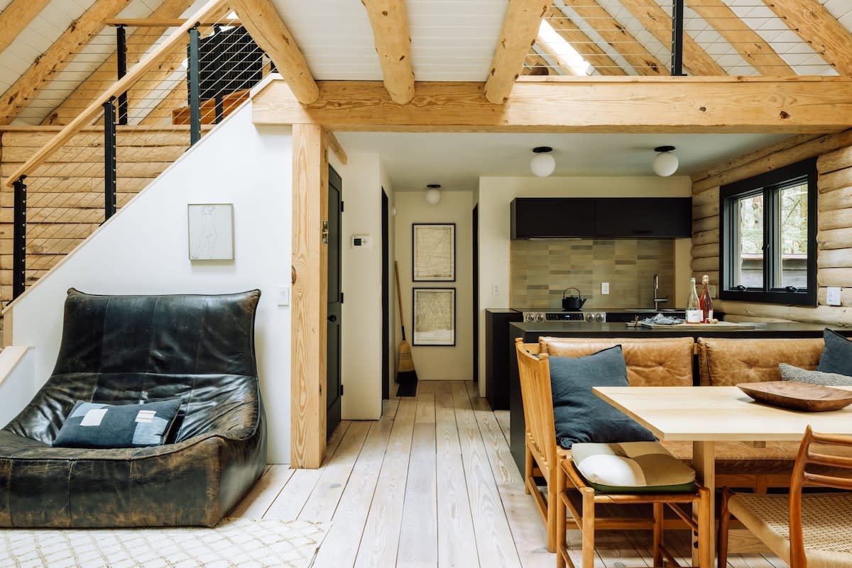 A wide view of the open-concept main floor shows a distressed leather sofa near the entrance, leading to a dining area with a leather bench and a modern kitchen beyond. The space features log walls, light wood floors, and a staircase to a loft.