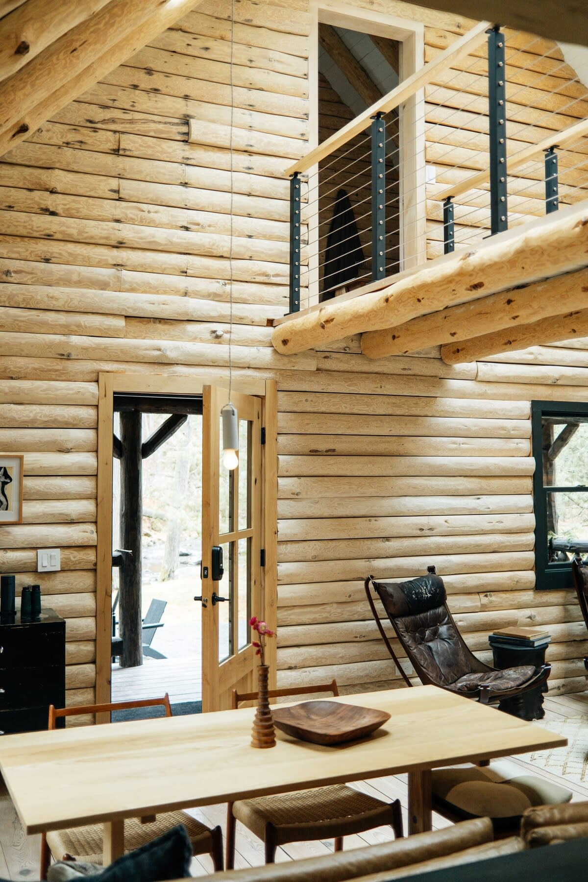 The bright interior of a log cabin features light-colored log walls and exposed log beams, creating a warm, natural aesthetic. A wooden dining table and a leather lounge chair define the open-concept living and dining space, overlooked by a loft with a metal railing.