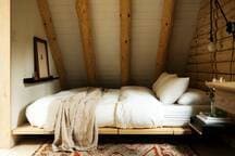 A cozy bedroom in the loft area features a low-profile bed with crisp white bedding, set against a light-colored log wall. The room boasts a sloped ceiling with exposed log beams and a small wall shelf adorned with a framed picture.