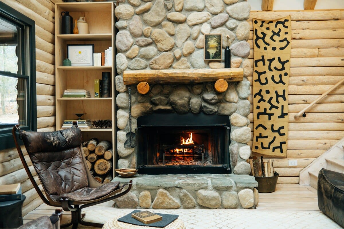A warm living room centers around a large stone fireplace with a burning fire, flanked by a built-in wooden bookshelf and a comfortable leather armchair. Log walls and light wood floors create a rustic yet inviting atmosphere.