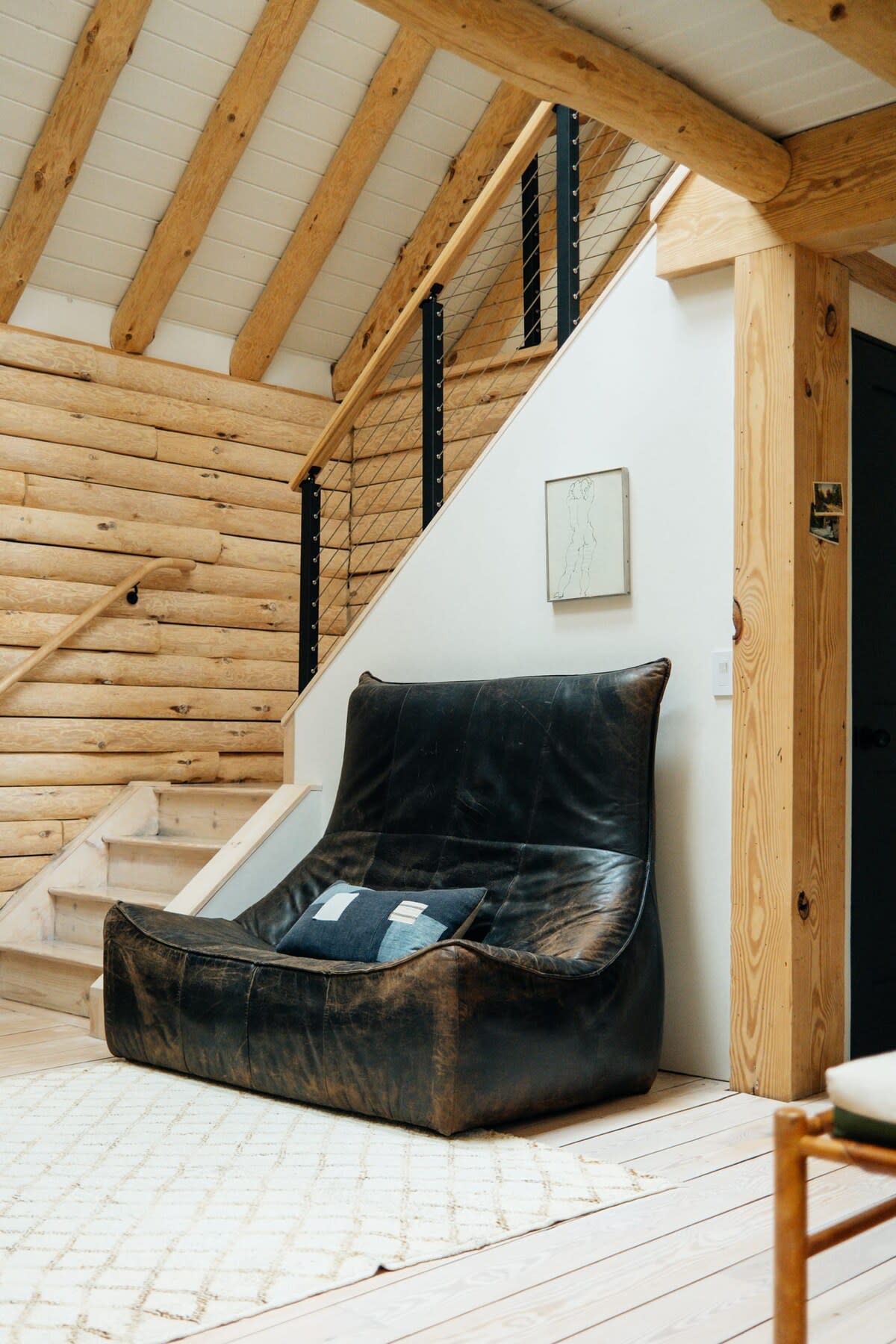 A spacious living area features a large, distressed leather sofa with a blue accent pillow, positioned beneath a modern staircase with black metal railings. The room has light wood floors, log walls, and a white accent wall.