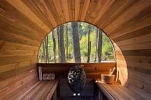 Interior of a barrel-shaped wooden sauna with benches on either side and a large arched window offering a view of the surrounding forest. A sauna heater and bucket are visible.