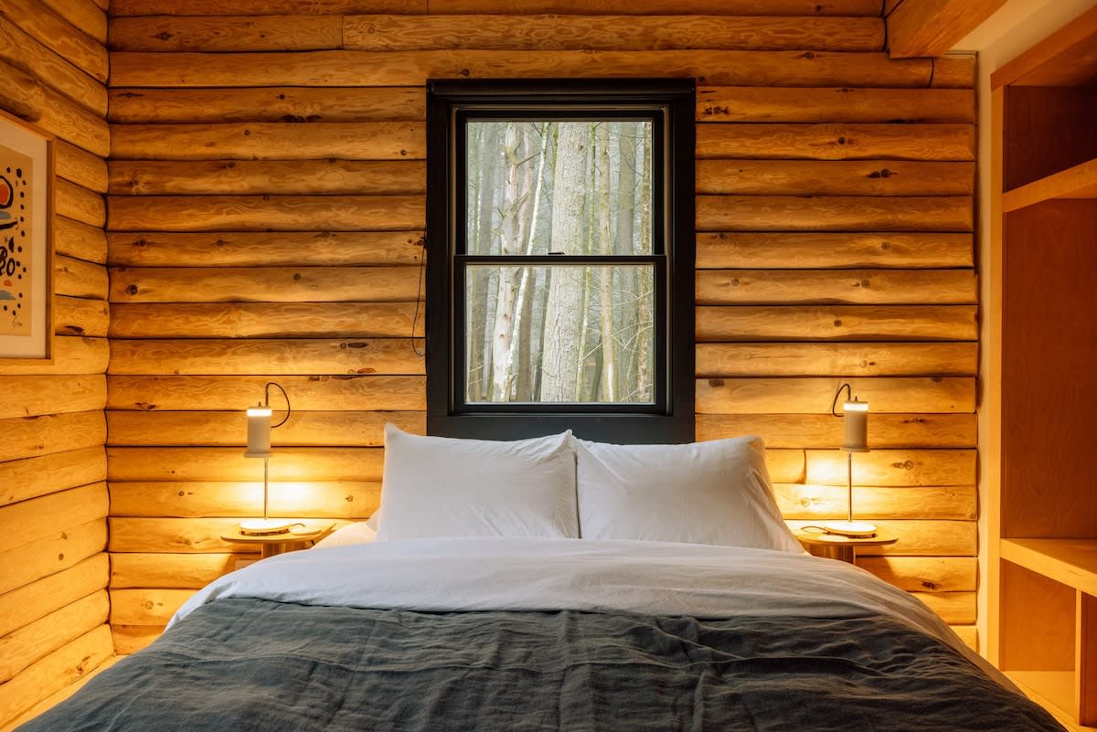 Inviting bedroom with horizontal log walls and a large bed with white bedding and a dark grey throw. Two modern bedside lamps illuminate small wooden nightstands, flanking a black-framed window offering a view of trees.