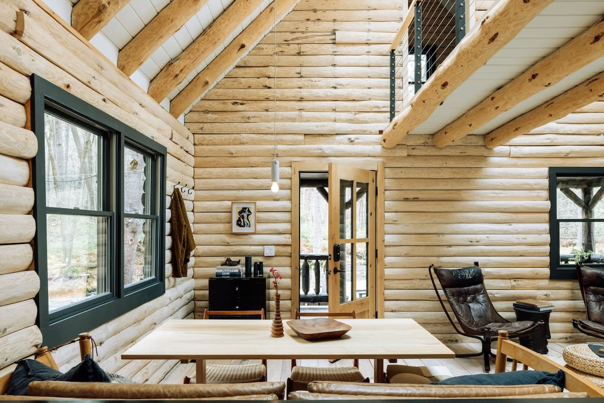 An open-concept living and dining area features natural log walls and exposed wooden beams, with a light wood dining table and chairs, and a glimpse of a leather lounge chair.