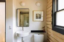 A bright bathroom with a white floating sink, black-framed rectangular mirror, and a toilet. The walls are a mix of light paint and natural wood planks, with a black shelf above the toilet and a window providing natural light.