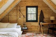 A rustic bedroom in a log cabin loft, featuring a low-profile bed with white bedding, light log walls, and a sloped ceiling with exposed beams. A dark-trimmed window overlooks the forest, and a wooden desk with a chair provides a workspace.