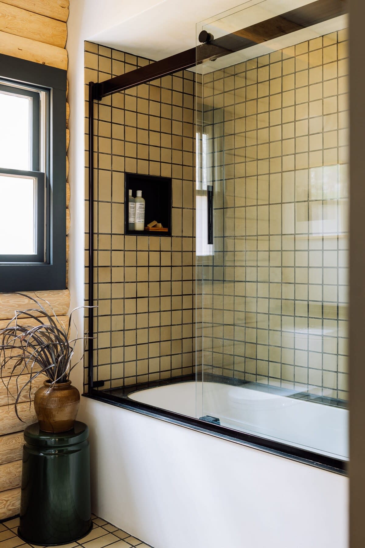Contemporary bathroom featuring a bathtub with a glass shower screen and light-colored square tiles with dark grout. A built-in shower niche holds toiletries, and a decorative plant sits on a dark green stool.