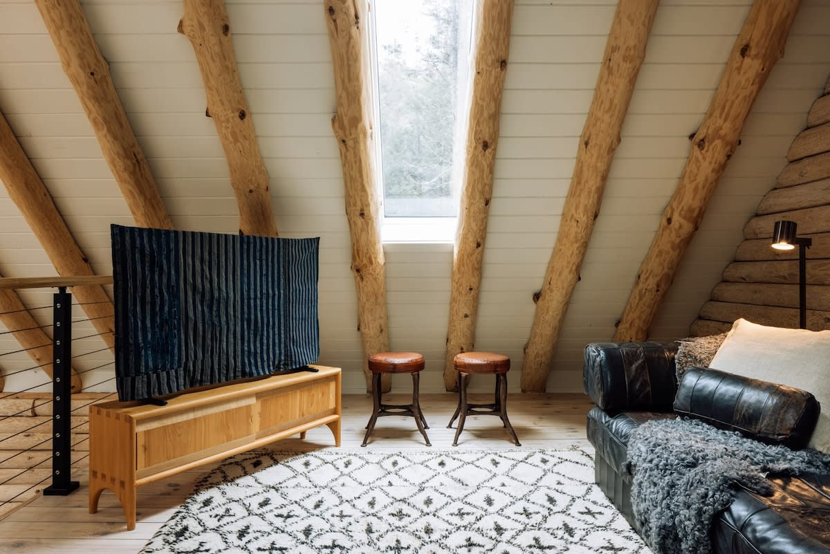 Cozy living area with a sloped, white-paneled ceiling and exposed log beams. Features a large TV on a wooden console, a dark leather sofa with a throw, two leather stools, and a patterned rug. A large skylight window brightens the space.