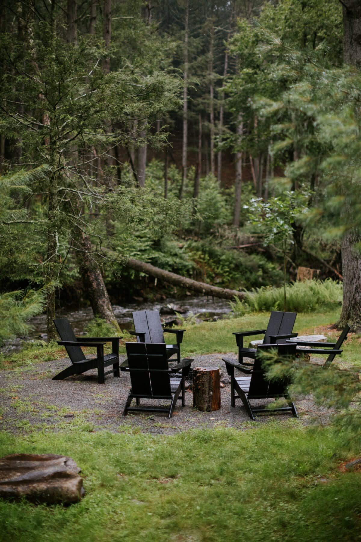 An inviting outdoor fire pit area with black Adirondack chairs arranged around a tree stump table, set on a gravel patch amidst a lush green forest with a river in the background.
