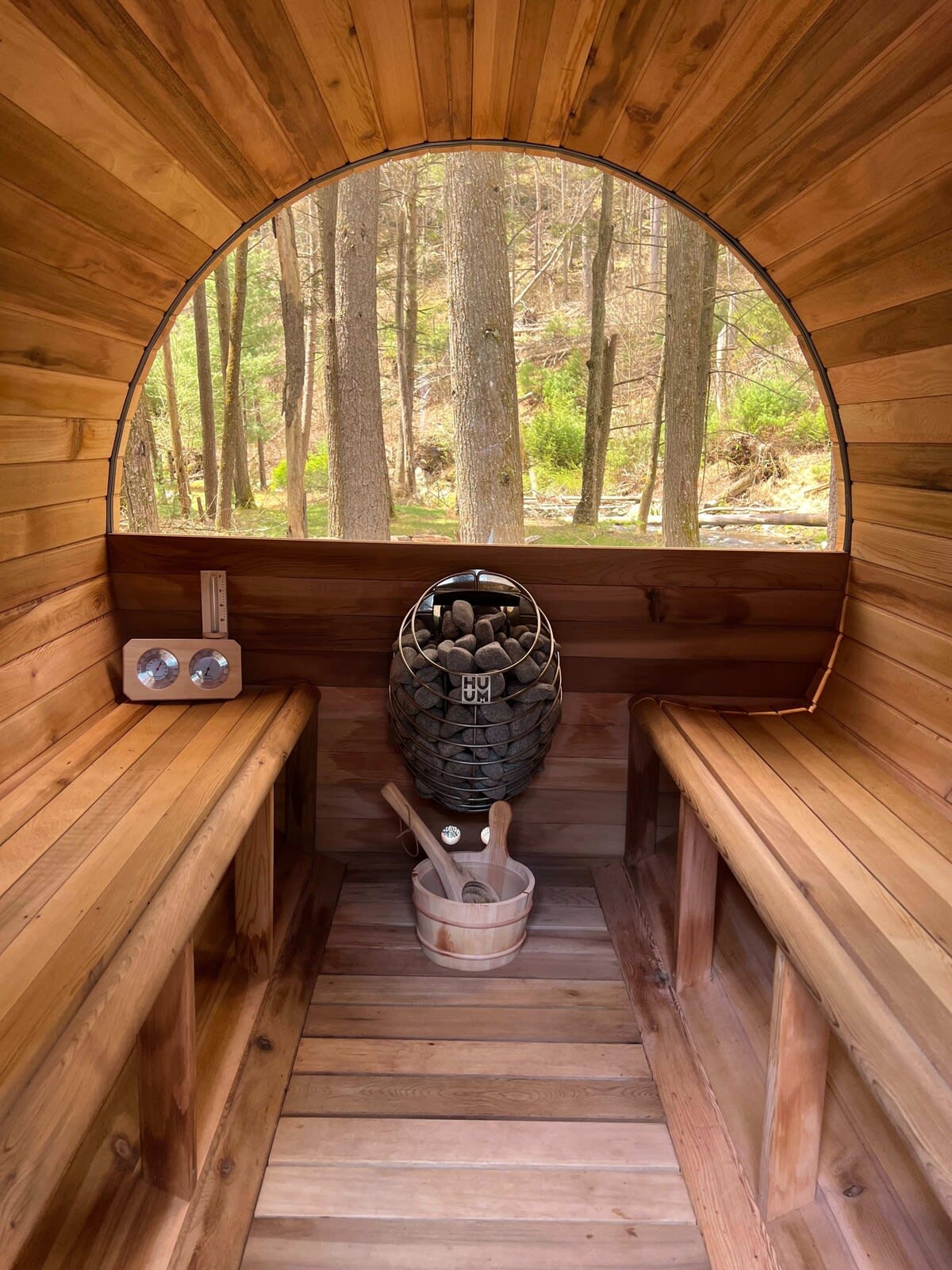 Interior of a barrel sauna with light wood paneling, two benches, a stone heater, and a large arched window offering a view of a dense forest. A wooden bucket and ladle are on the floor.