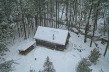 An aerial view of a rustic log cabin and a smaller outbuilding, both with snow-covered roofs, situated in a dense, snow-laden forest. Tracks in the snow lead to the main cabin.