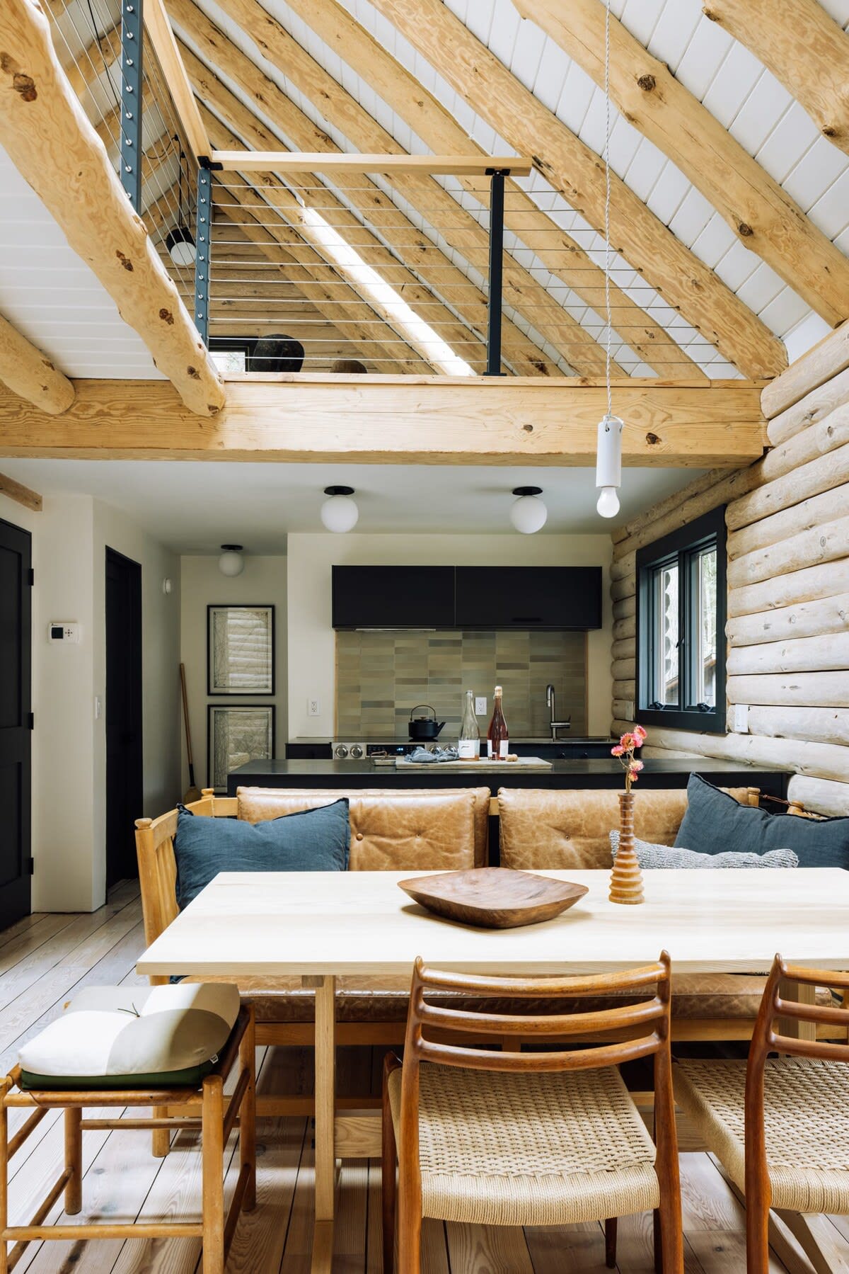 An open-concept living space features a dining area with a light wood table and leather bench seating, flowing into a modern kitchen with dark cabinetry and a tiled backsplash. Exposed log beams and a loft area with metal railings are visible above.