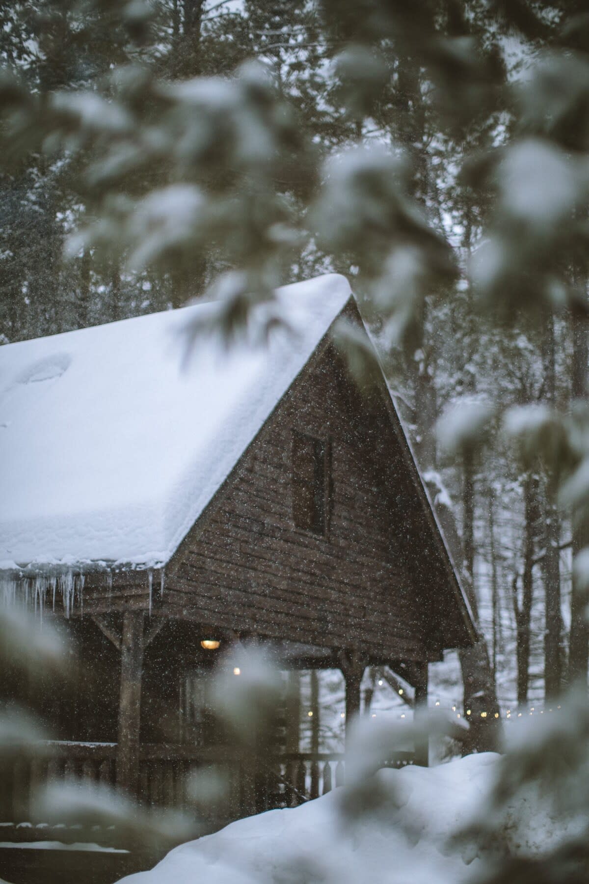 Close-up view of the log cabin's snow-covered roof and side, with icicles hanging from the eaves, seen through blurred snowy tree branches. A warm light glows from the porch.