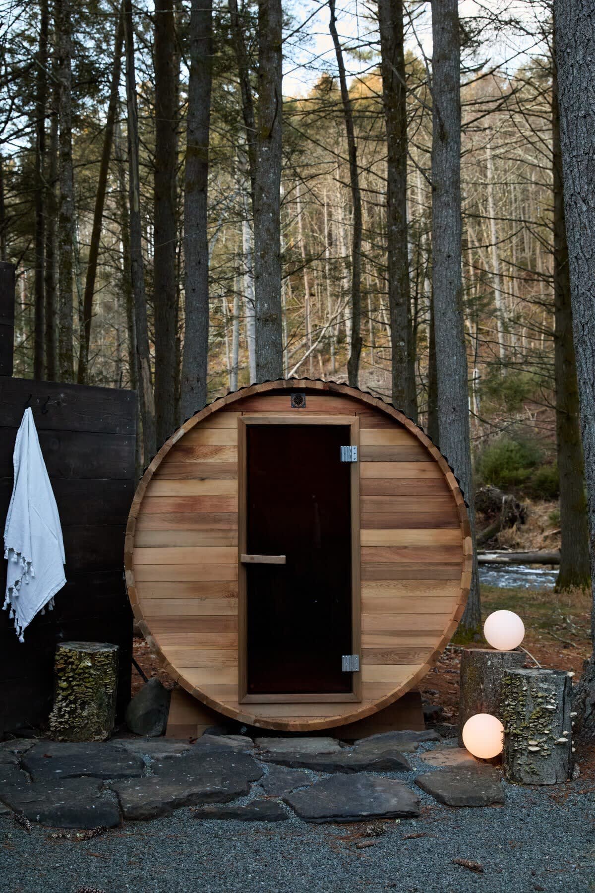 Exterior of a wooden barrel sauna with a dark glass door, situated on a stone and gravel base in a forest setting. Two glowing orb lights and a white towel hang nearby.