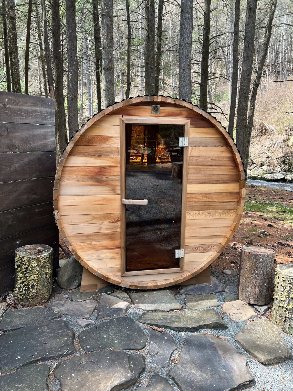 Outdoor barrel sauna made of light-colored wood, featuring a dark glass door and set on a stone patio. It is surrounded by tall trees, suggesting a secluded, natural setting.