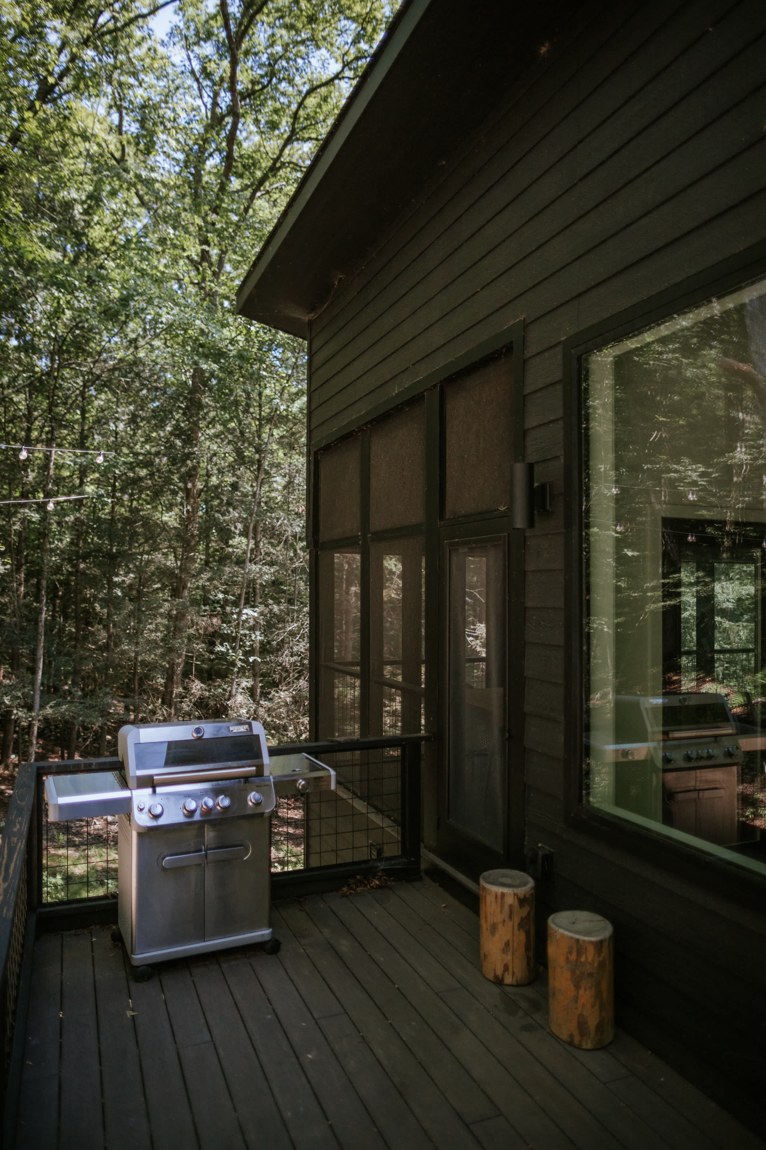 A dark-stained wooden deck features a stainless steel BBQ grill and two rustic log stools, set against a backdrop of dense green forest and a dark-colored modern house exterior.