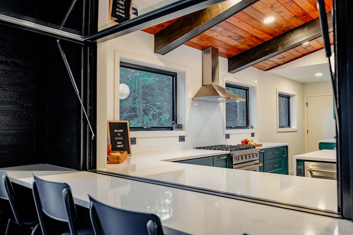 The kitchen is viewed through a large pass-through window, revealing dark green cabinetry, white subway tile backsplash, and a stainless steel range hood. A 'Welcome to Little Rock Cabin' sign adds a personal touch, with forest views visible through the windows.