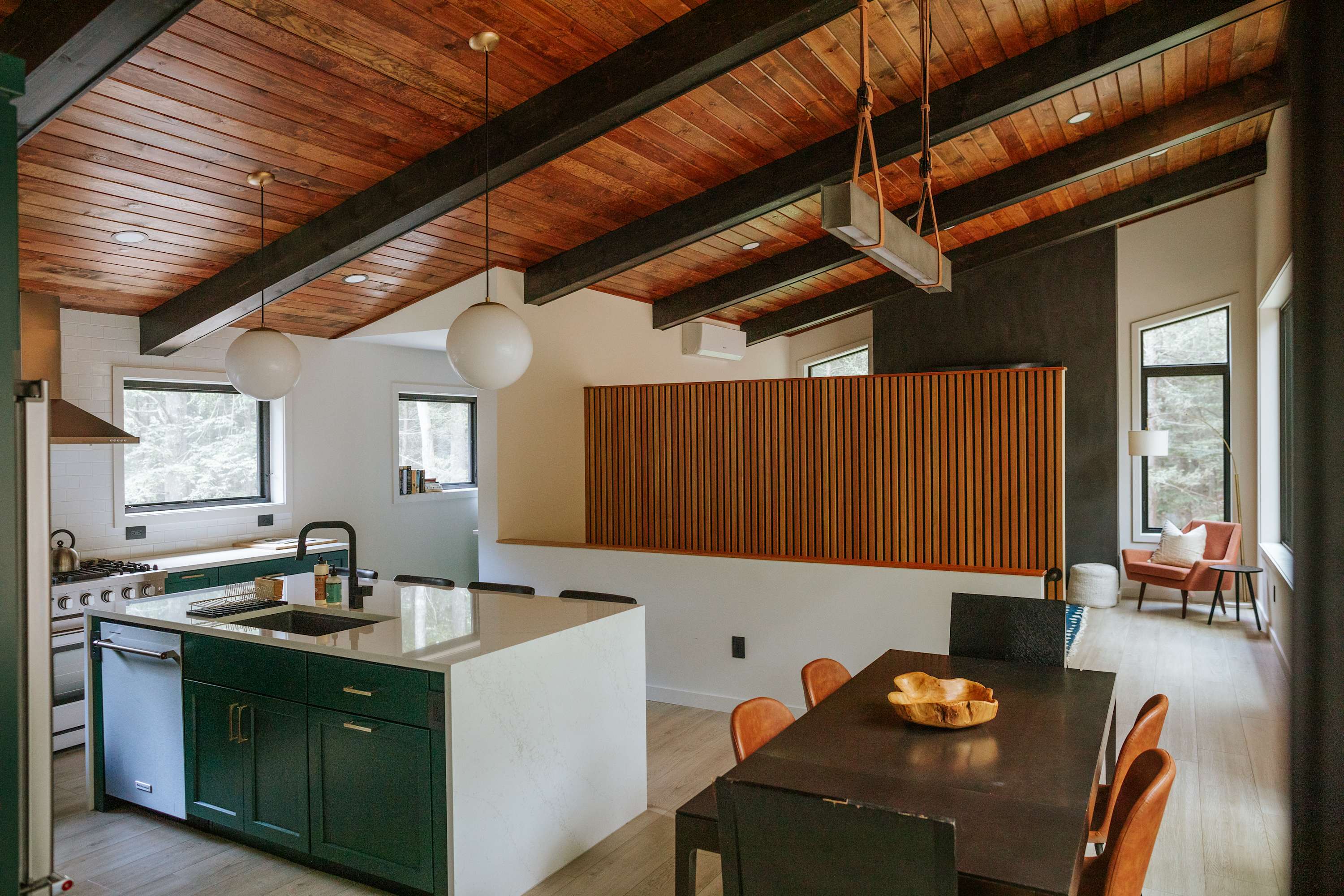 Open-concept kitchen and dining area featuring dark green cabinetry, a white island with bar seating, and a dark dining table with orange chairs. The space has a striking wooden sloped ceiling with dark beams and a decorative wooden slatted wall.