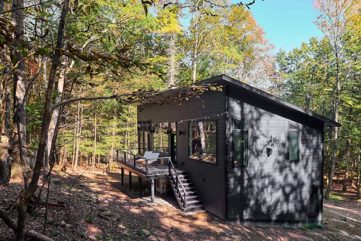 Modern dark-sided house with a sloped roof and large windows, featuring a wooden deck with outdoor seating, surrounded by a sunlit forest.
