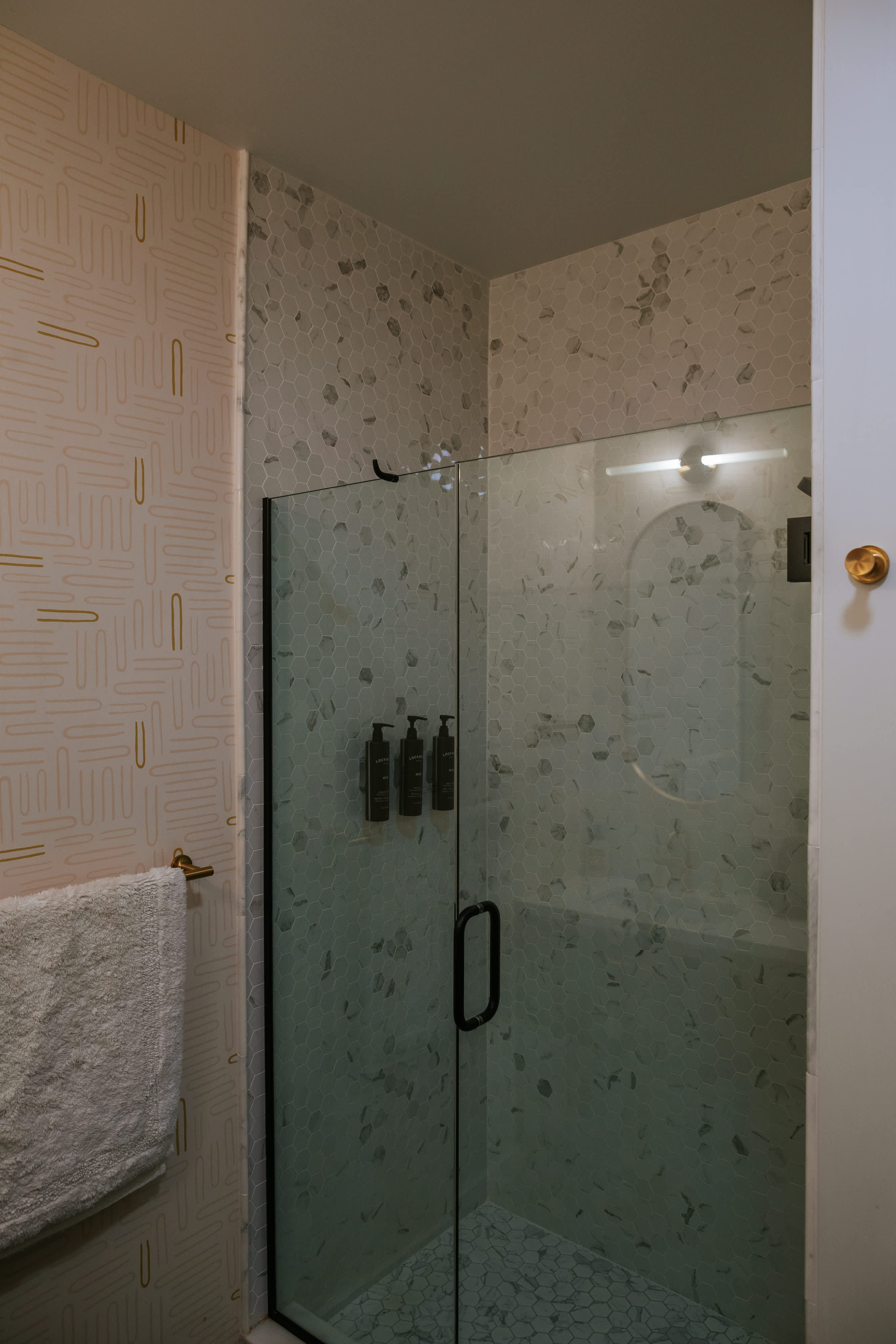 A modern shower enclosure with a glass door and black frame is visible, featuring light grey hexagonal tiles on the walls and floor, adjacent to a wall with light pink and gold paperclip-patterned wallpaper.