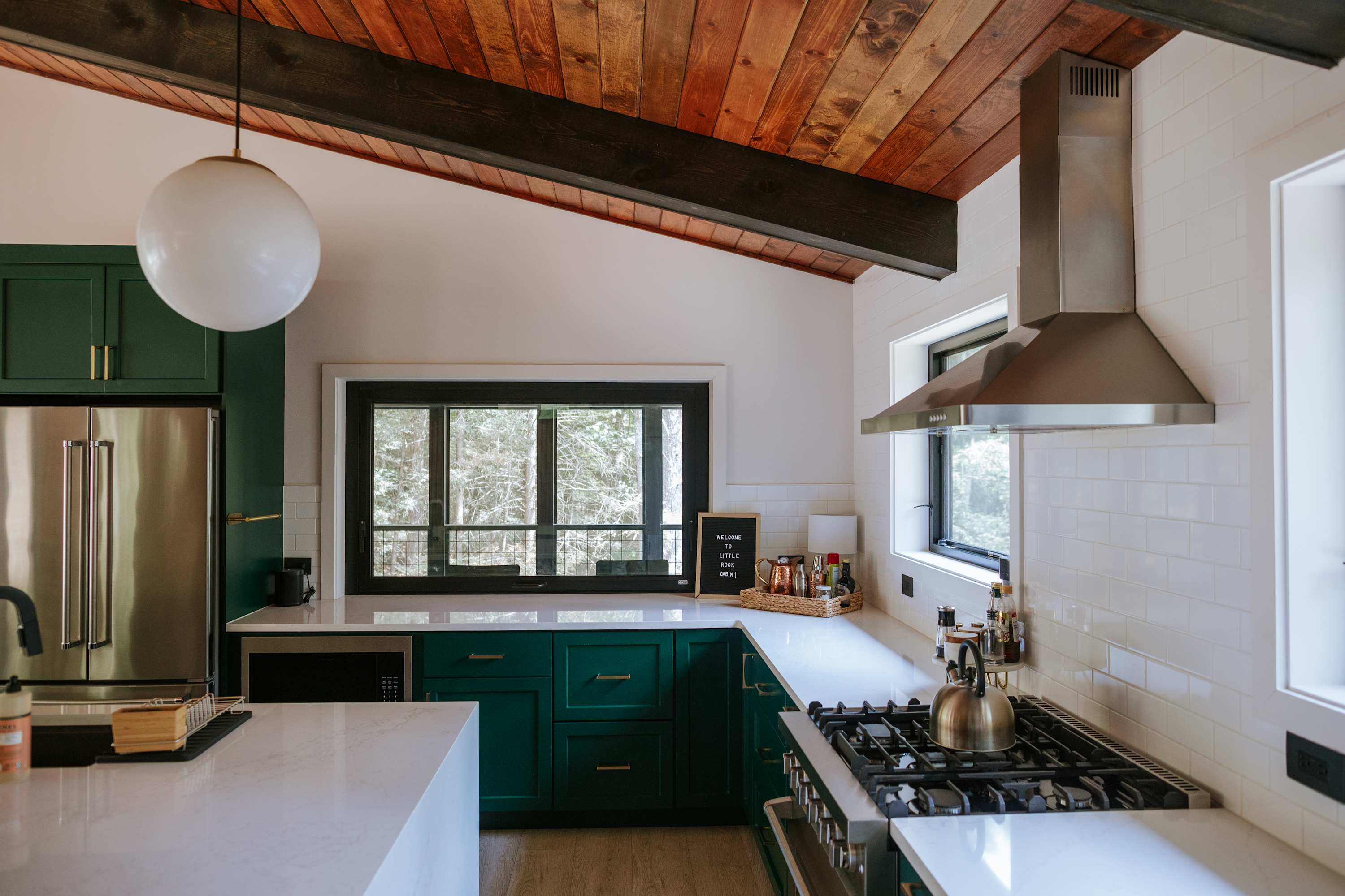 Bright kitchen with dark green lower cabinets, white subway tile backsplash, and white quartz countertops. A stainless steel range hood hangs over a gas stove, and large windows provide a view of the surrounding trees.