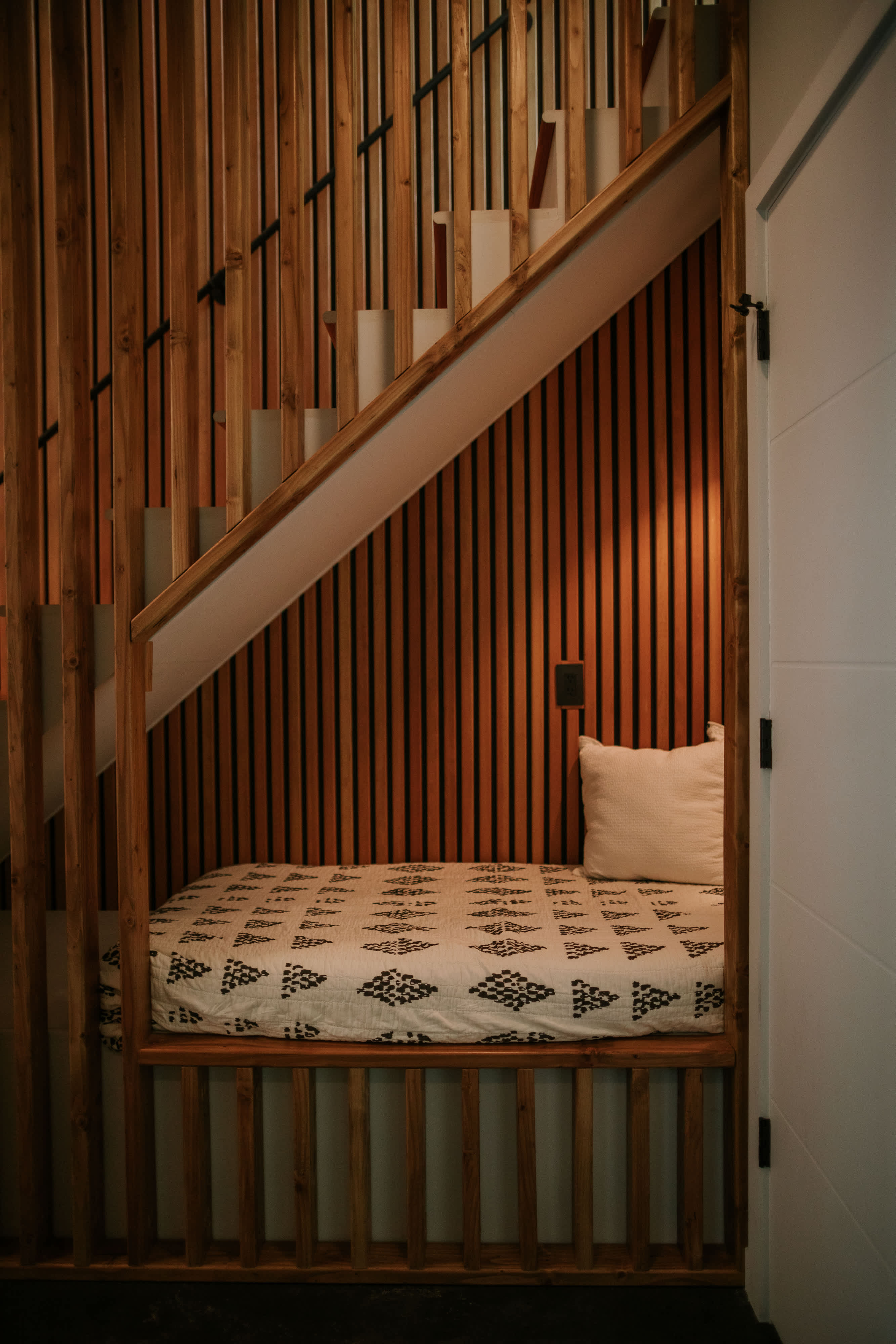 A cozy built-in nook under a wooden slatted staircase features a patterned mattress and a white pillow, surrounded by warm wooden paneling and vertical slats, creating a private sleeping or lounging area.