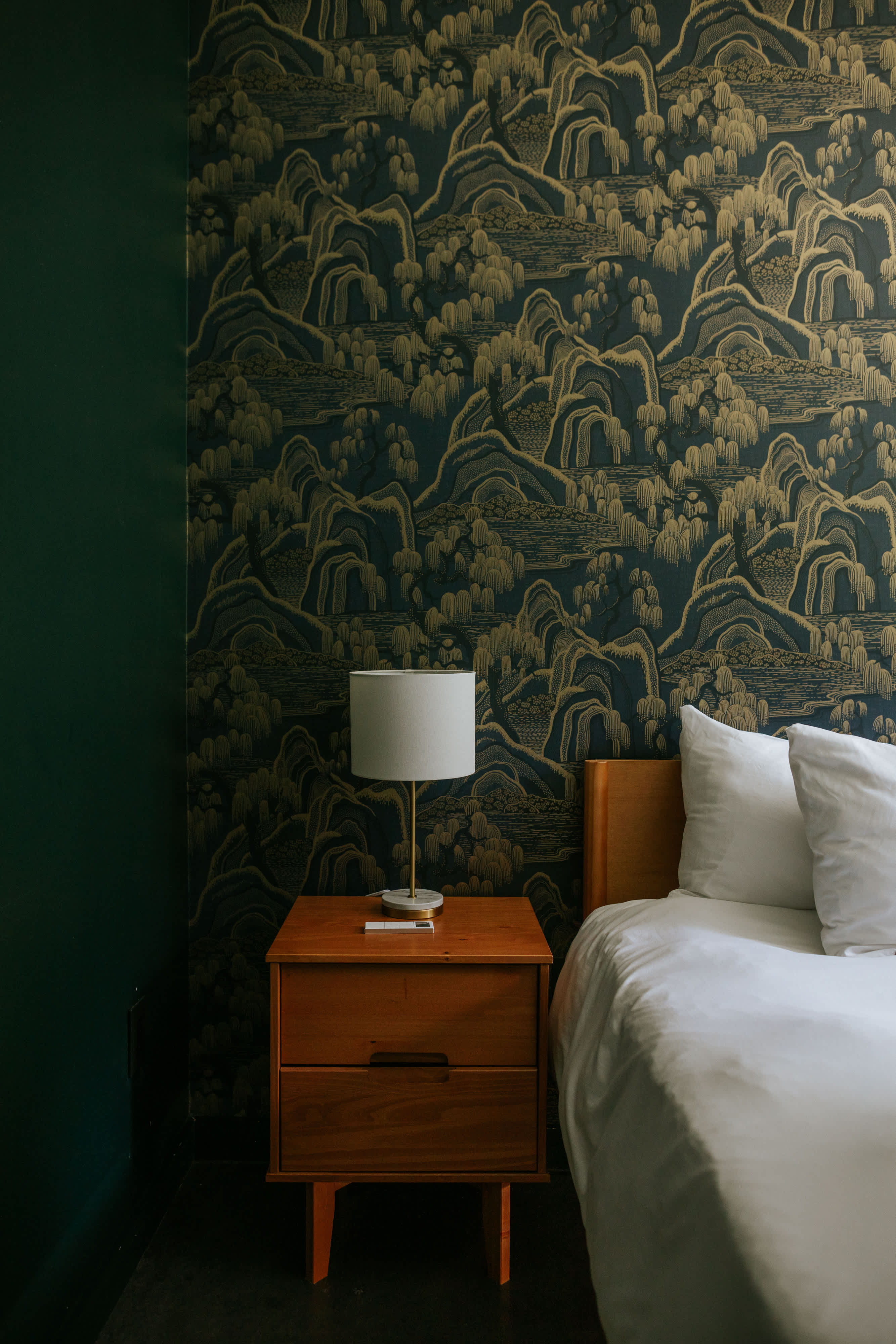 A close-up of a bedroom corner shows a wooden nightstand with a modern lamp next to a bed with white linens, against a wall adorned with dark blue and gold scenic wallpaper, complementing the dark green wall.