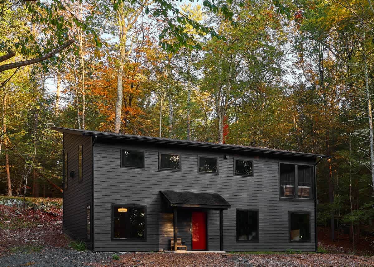 Modern dark gray house with horizontal siding, nestled among vibrant autumn trees, featuring a bright red front door. The two-story structure has multiple dark-framed windows and a small covered entrance.